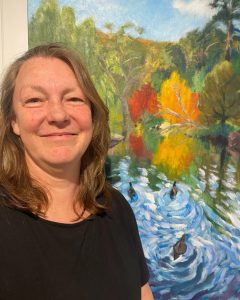 Jodie Kilmister standing in front of her landscape painting featuring water and ducks in a vibrant natural setting