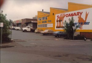 Historic photograph of Springett Wholesale Pty Ltd in Bowral, featuring the bright yellow warehouse, Hot Canary signage and vintage vehicles.